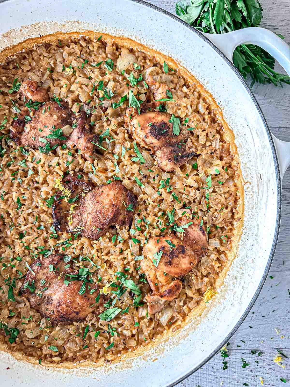 A large white Dutch oven filled with browned chicken thighs cooked on top of creamy rice, all garnished with chopped fresh parsley. A bunch of fresh parsley sits nearby on the light wooden surface.