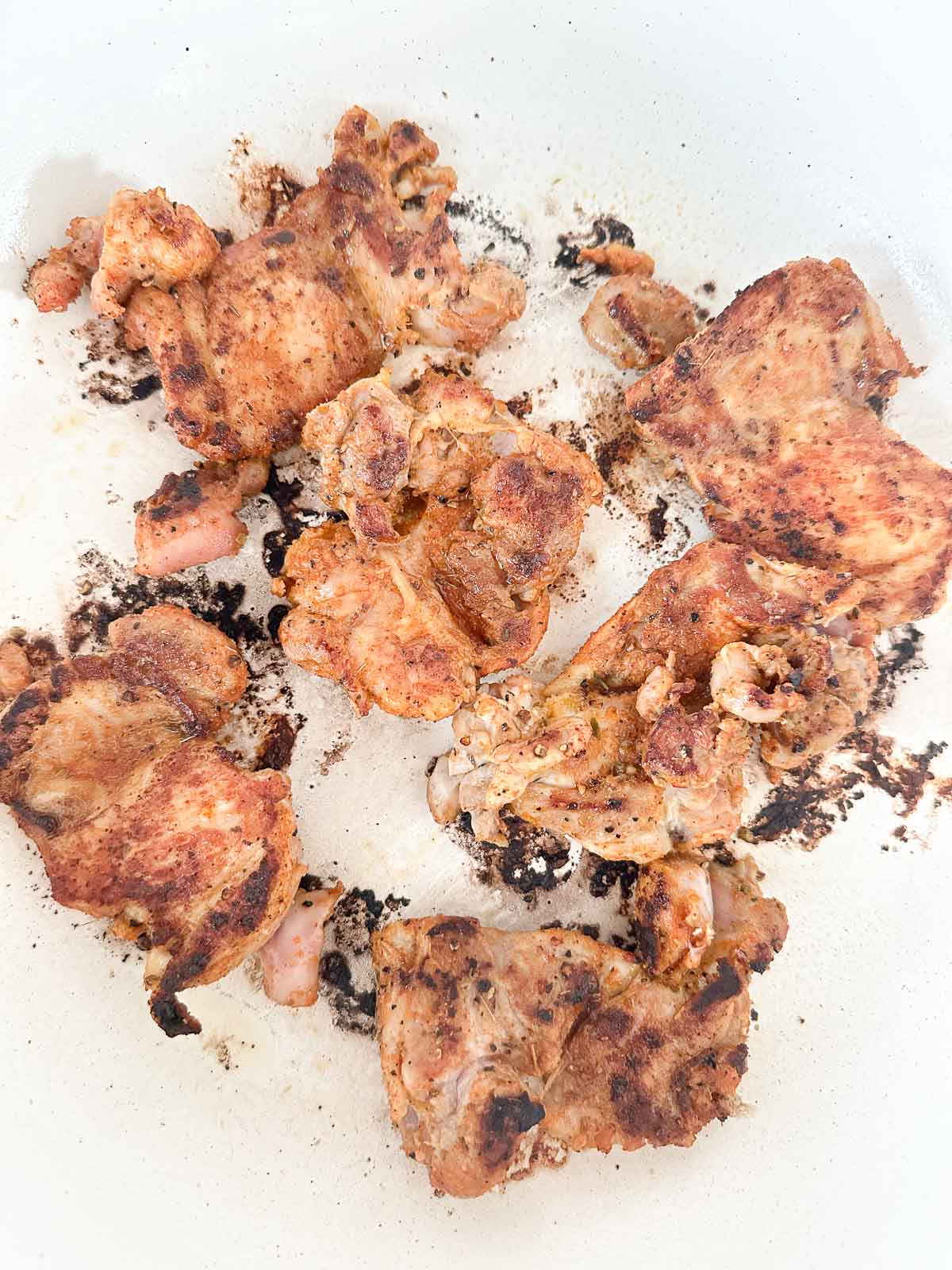 Several pieces of seasoned, cooked chicken are scattered in a white pan, with some charring and browned spots visible on both the chicken and pan surface.