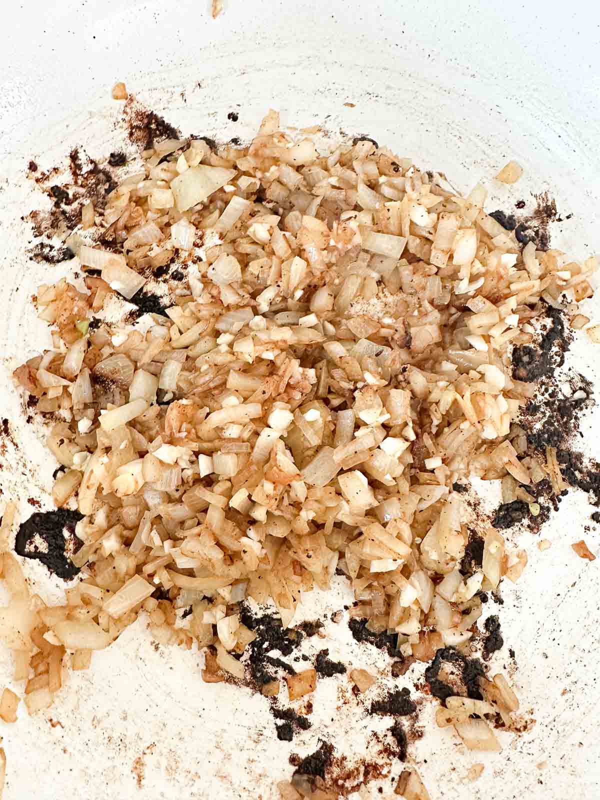 Chopped onions sautéing in a white pan, turning golden brown with some charred bits sticking to the pan.