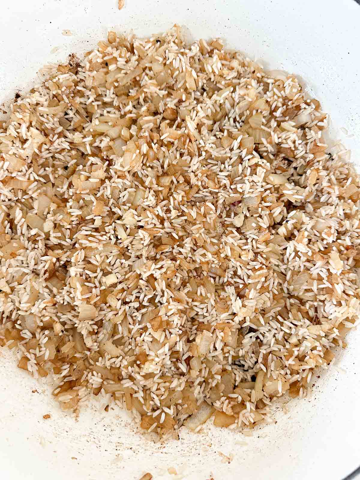 A close-up of uncooked rice, chopped onions, and shredded coconut being sautéed together in a white pan. The ingredients are lightly browned and evenly mixed.
