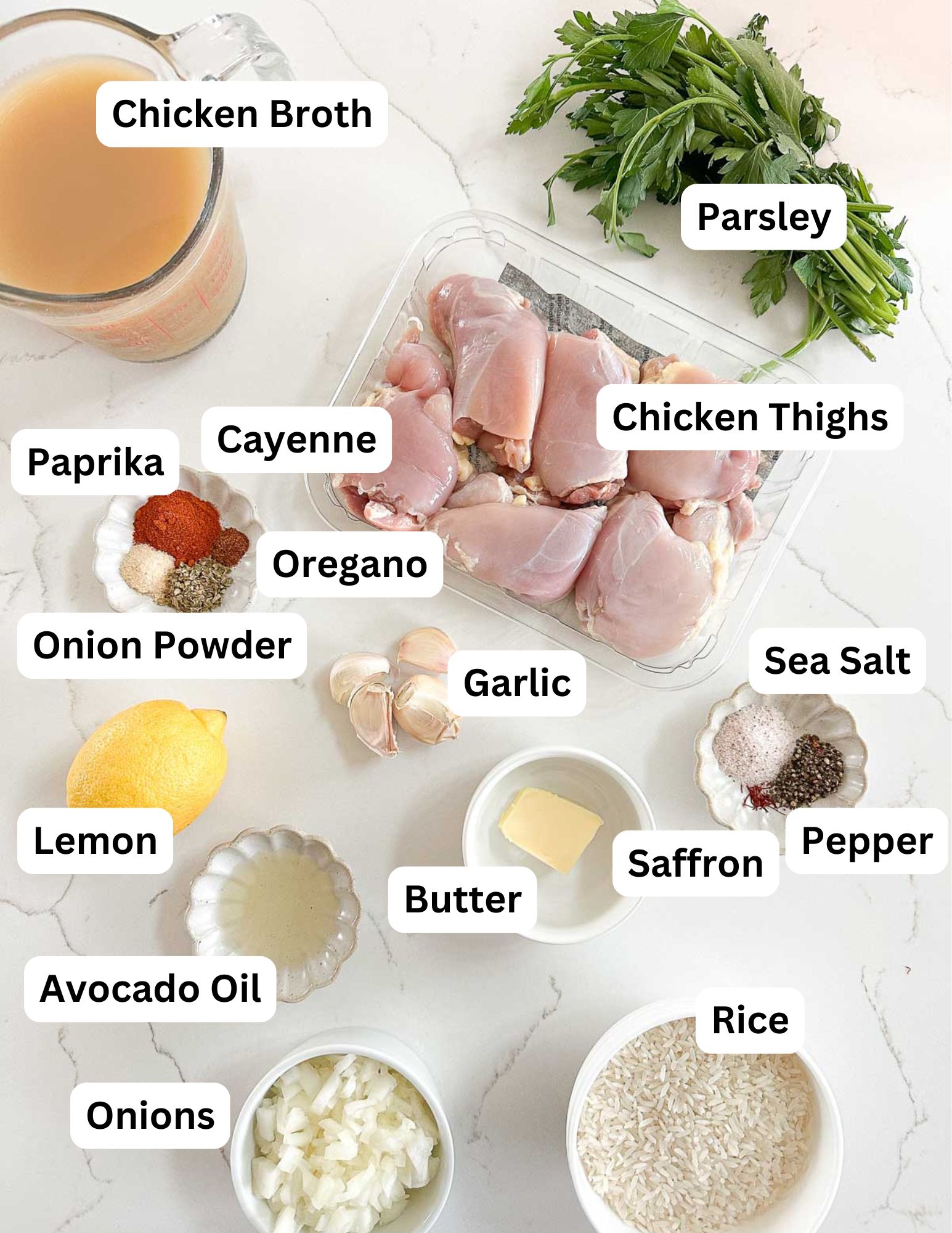 Top-down view of labeled ingredients on a white surface, including chicken broth, parsley, chicken thighs, paprika, cayenne, oregano, onion powder, garlic, sea salt, pepper, saffron, butter, rice, onions, lemon, and avocado oil.