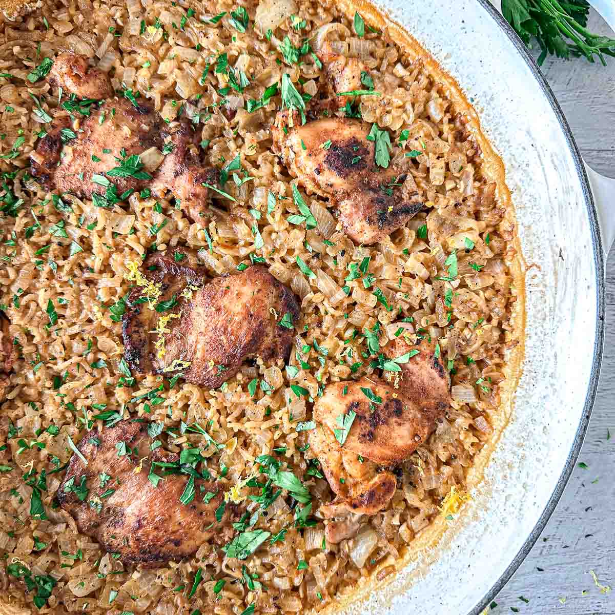 A skillet filled with seasoned chicken thighs on a bed of creamy rice, garnished with chopped fresh herbs.