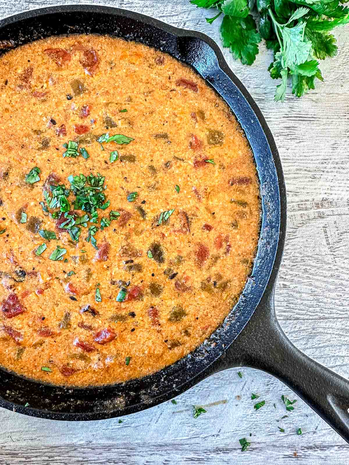 A cast-iron skillet filled with creamy, smoked queso dip garnished with chopped herbs sits on a light wooden surface. Fresh herbs are placed nearby.