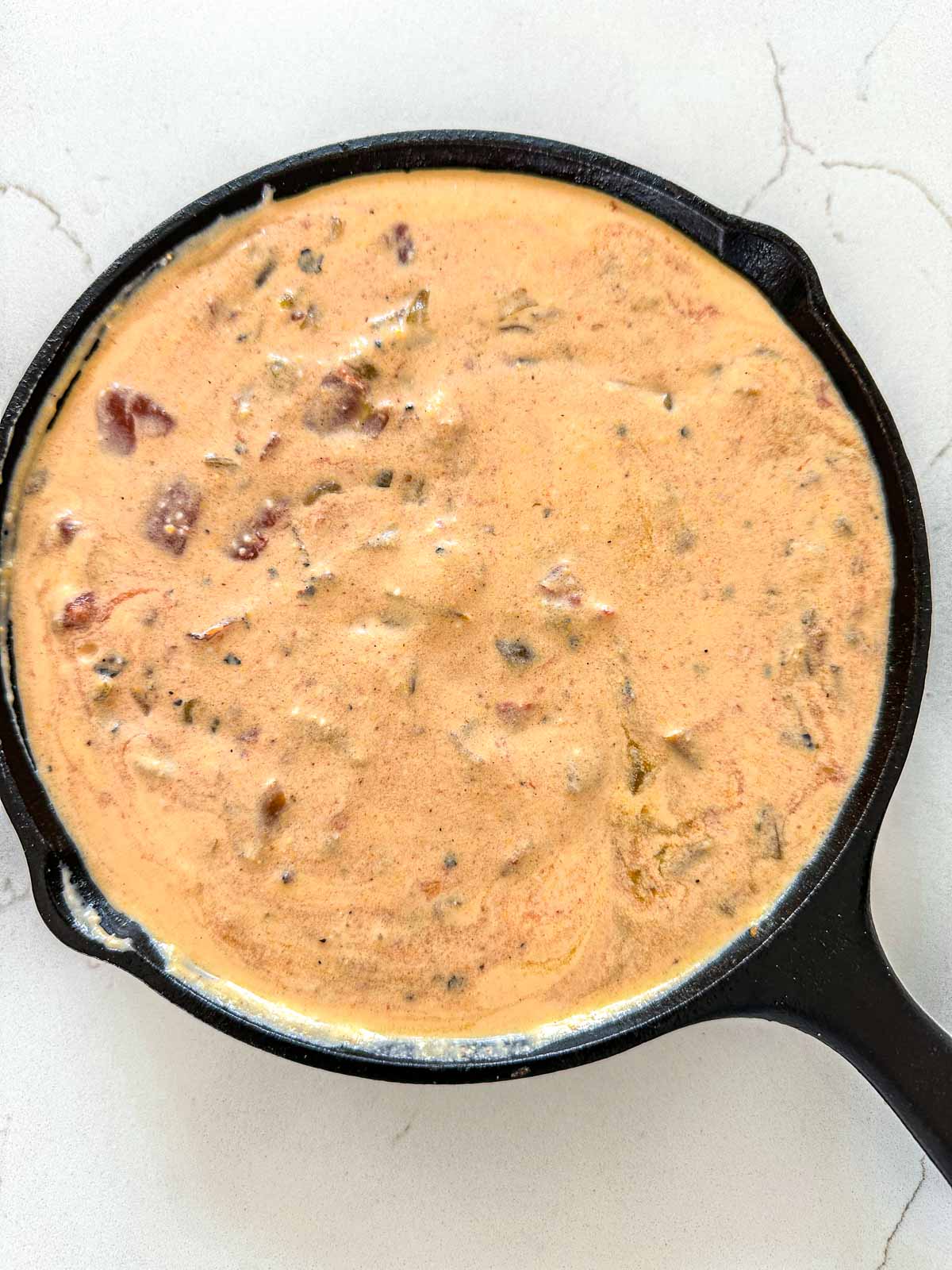 A cast iron skillet filled with smoked questo dip containing visible chunks of tomato and green chilies, sitting on a white countertop.