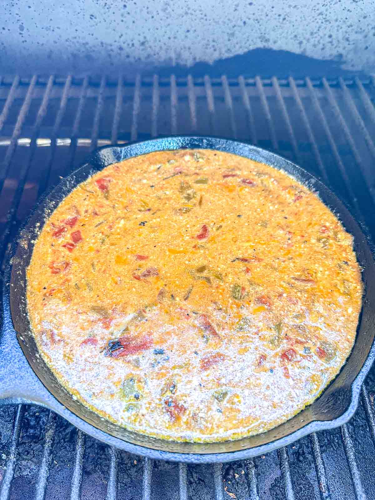 A cast iron skillet filled with a bubbling, cheesy dip containing tomatoes and green peppers sits on a grill with dark grates in the background.