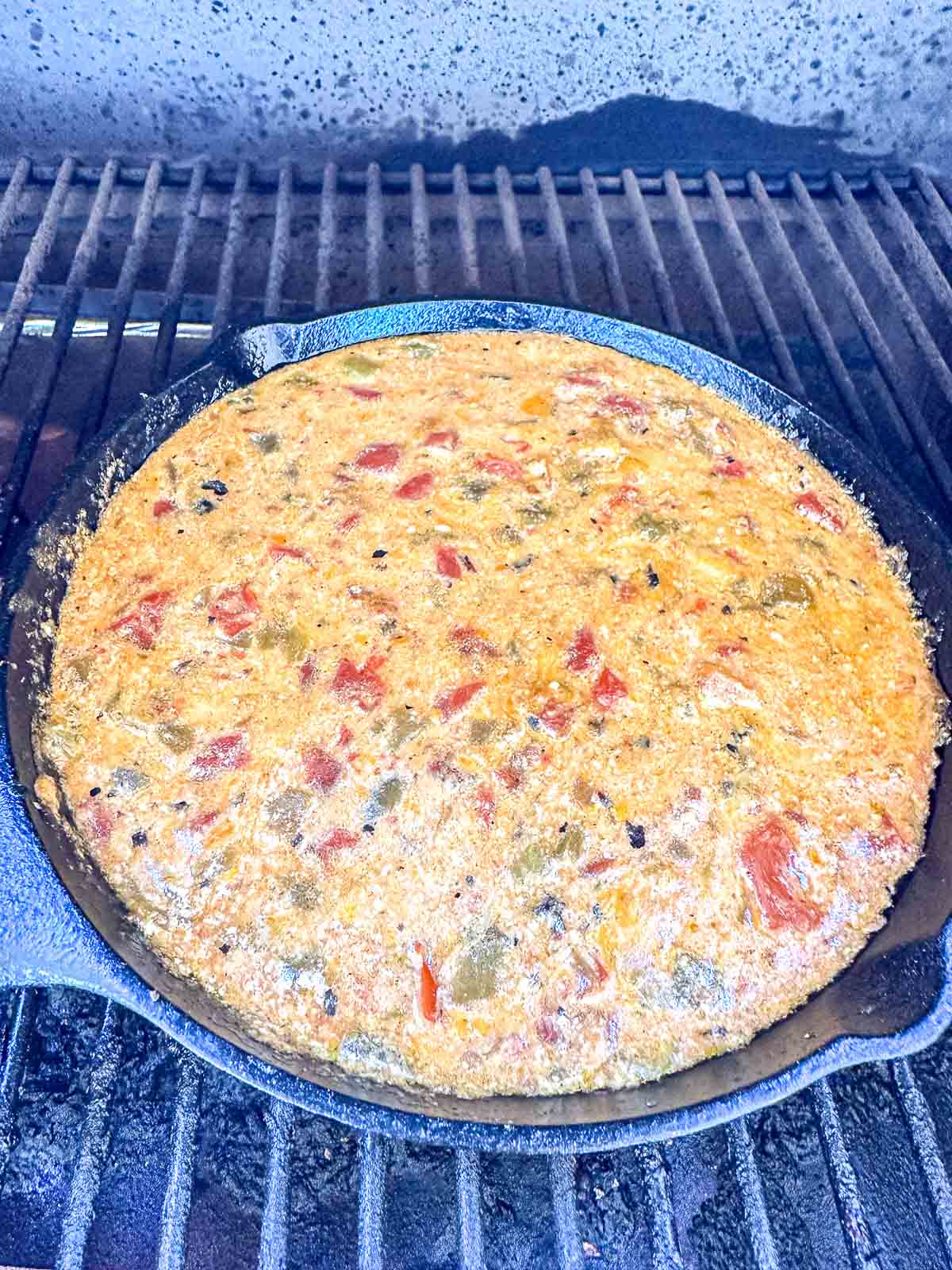 A cast iron skillet filled with smoked queso sits on the grates of an outdoor grill.