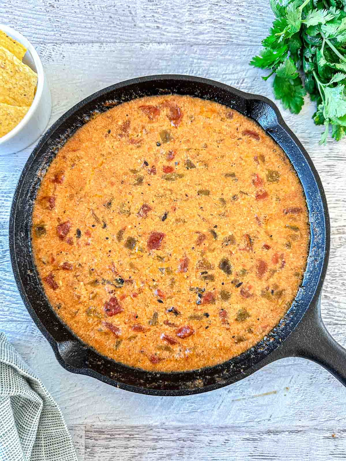A cast iron skillet filled with creamy, cheesy queso dip with tomatoes and peppers, set on a light wooden surface. Tortilla chips and fresh cilantro are nearby.