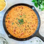 A cast-iron skillet filled with creamy, smoked queso dip topped with chopped herbs sits on a light wooden surface. Fresh cilantro and a bowl of tortilla chips are nearby.