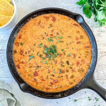 A cast-iron skillet filled with creamy, smoked queso dip topped with chopped herbs sits on a light wooden surface. Fresh cilantro and a bowl of tortilla chips are nearby.