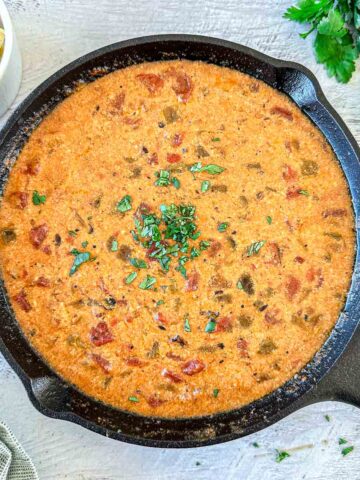 A cast-iron skillet filled with creamy, smoked queso dip topped with chopped herbs sits on a light wooden surface. Fresh cilantro and a bowl of tortilla chips are nearby.