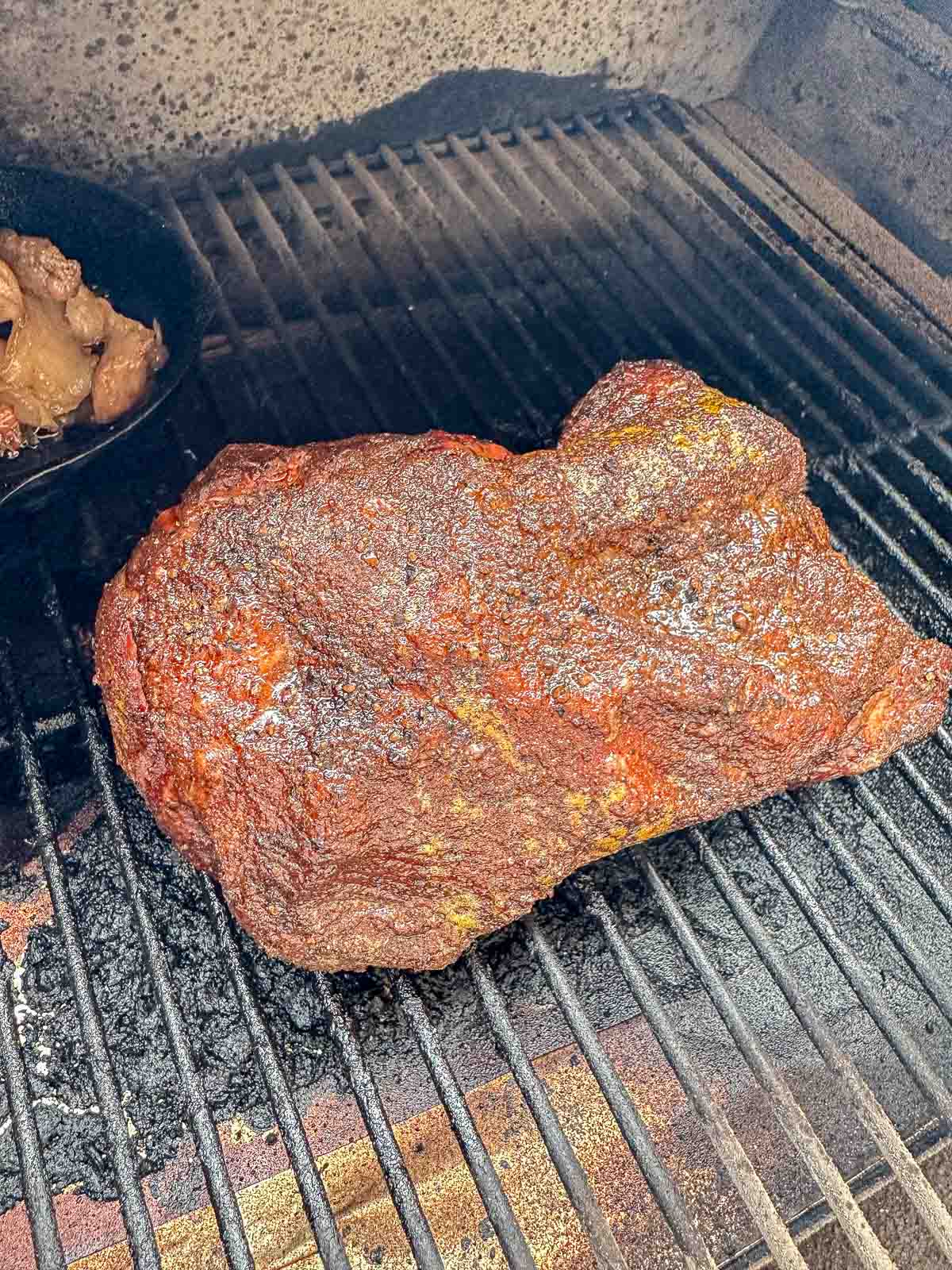 A large, seasoned piece of pork butt is smoking on a grill, developing a dark, crusty bark. In the background, a cast iron skillet sits on the grill beside the brisket.