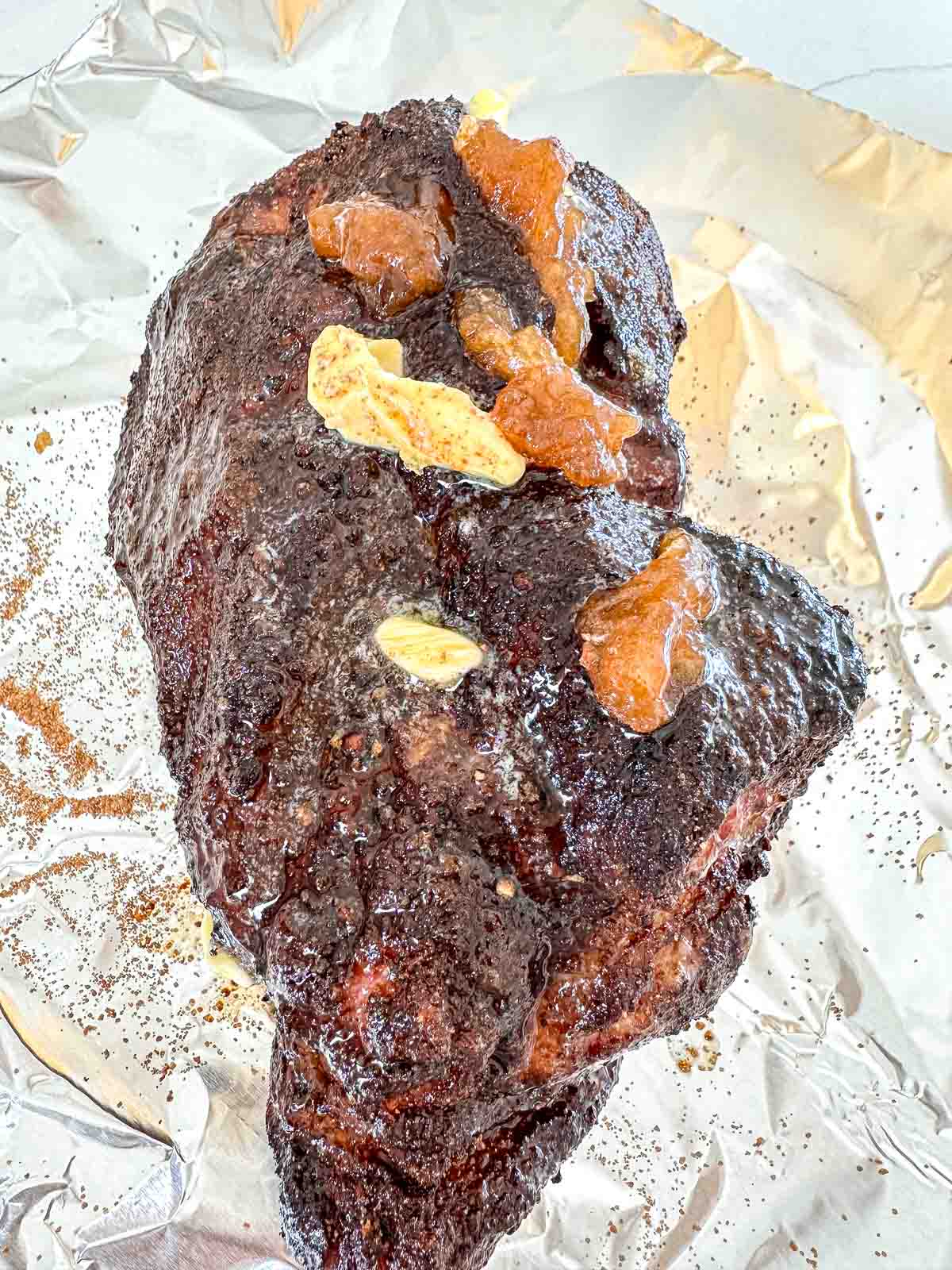 A large piece of seasoned, pork shoulder sits on foil, topped with several pats of butter and brown sugar. The meat appears dark and crusted, likely from a dry rub and slow cooking.