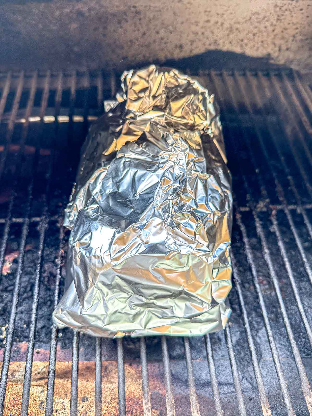 A large piece of pork shoulder wrapped in crinkled aluminum foil sits on the grates of a barbecue grill, ready to be cooked.