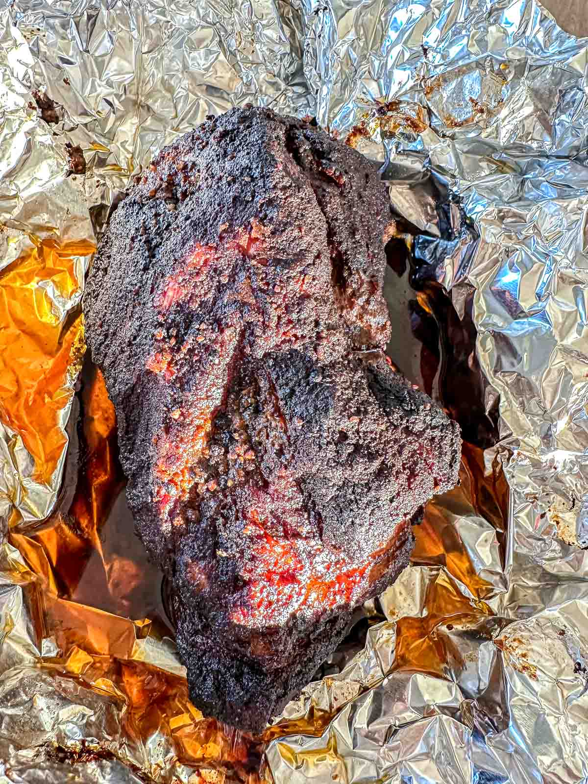 A large, darkly smoked piece of pork shoulder with a charred crust sits on shiny, crinkled aluminum foil, surrounded by juices.