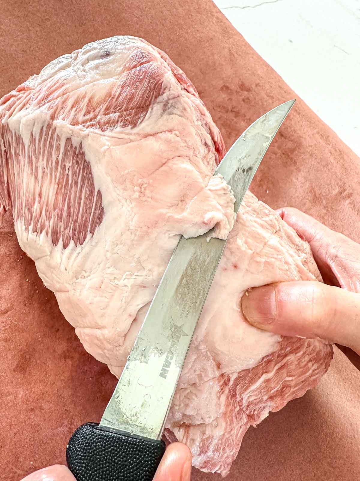 A person uses a knife to trim the fat off a raw piece of pork resting on brown butcher paper.