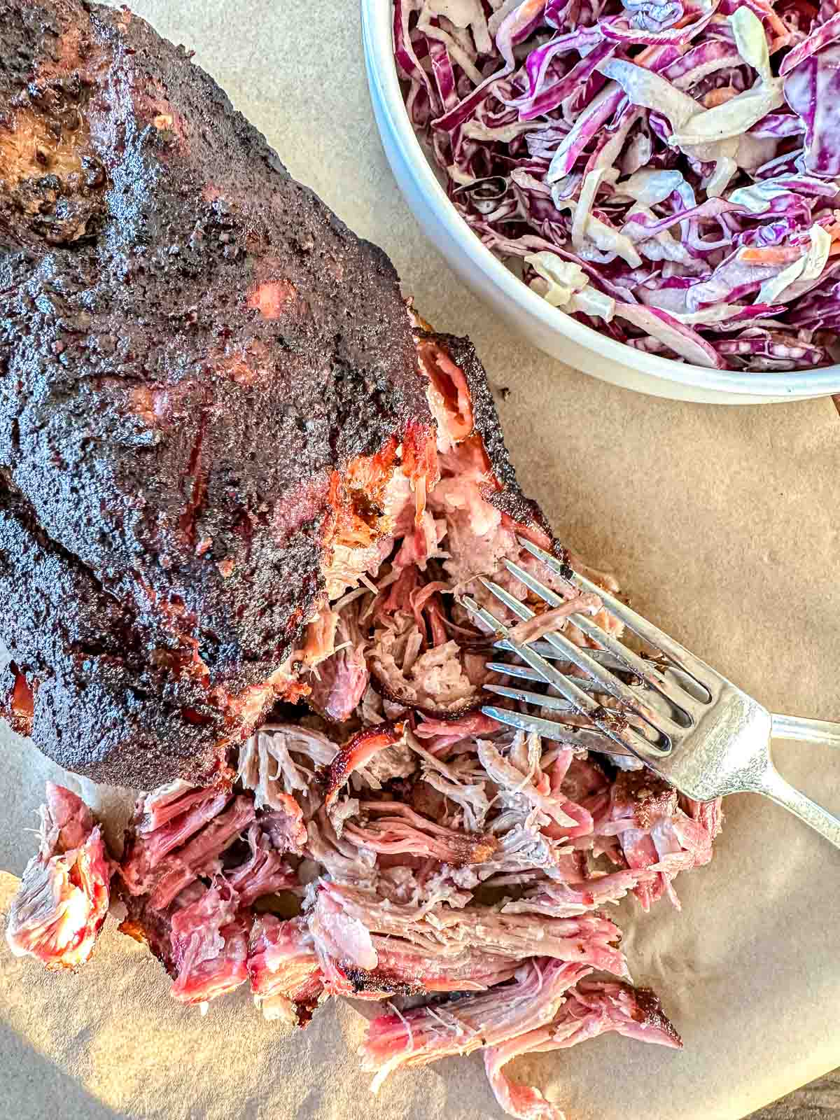 A close-up of smoked pulled pork being shredded with a fork on parchment paper, next to a bowl of coleslaw made with shredded red and white cabbage.