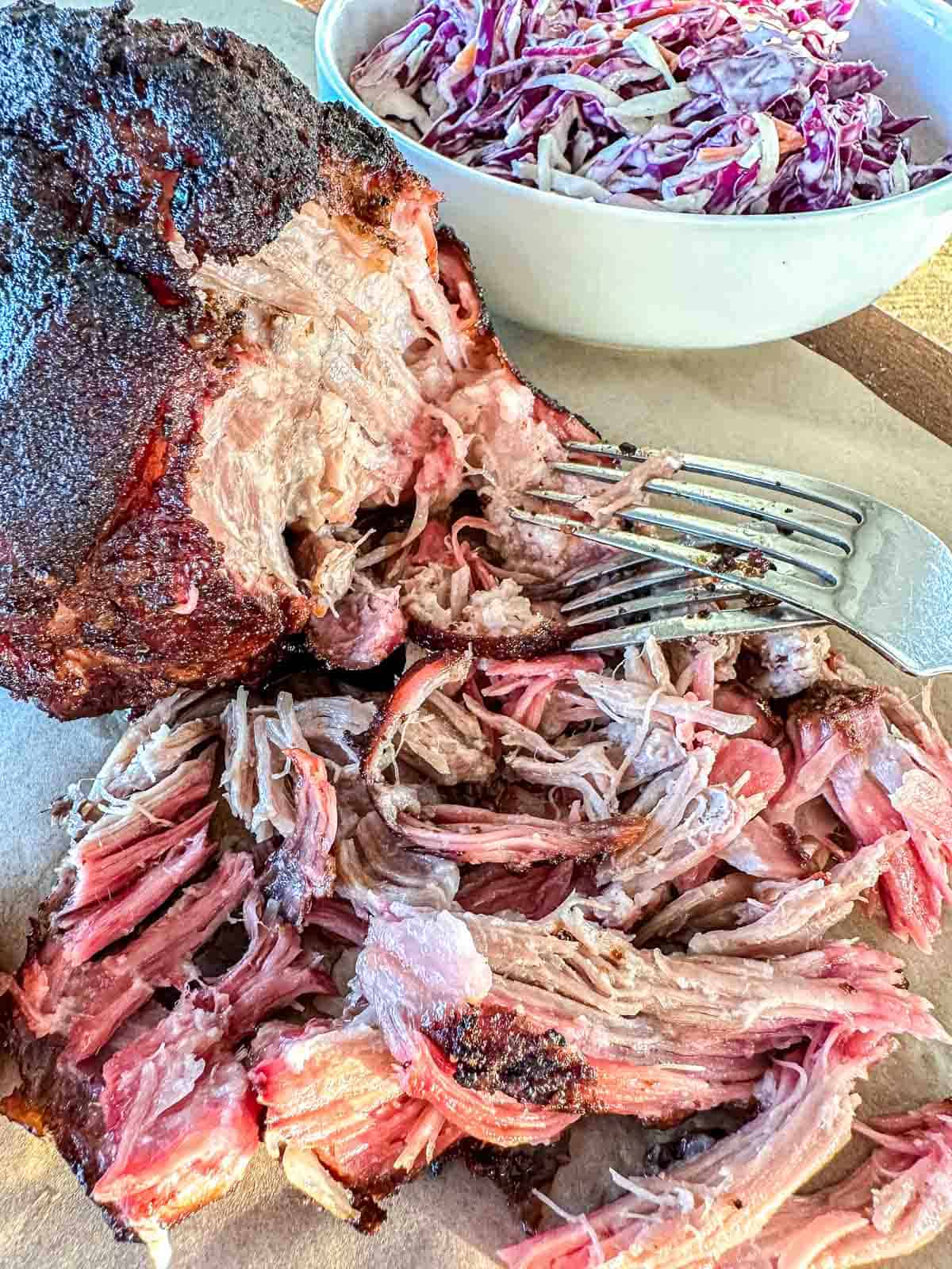 A close-up of shredded smoked pork butt with a fork next to a larger piece of pork, served on parchment paper, with a bowl of creamy coleslaw in the background.