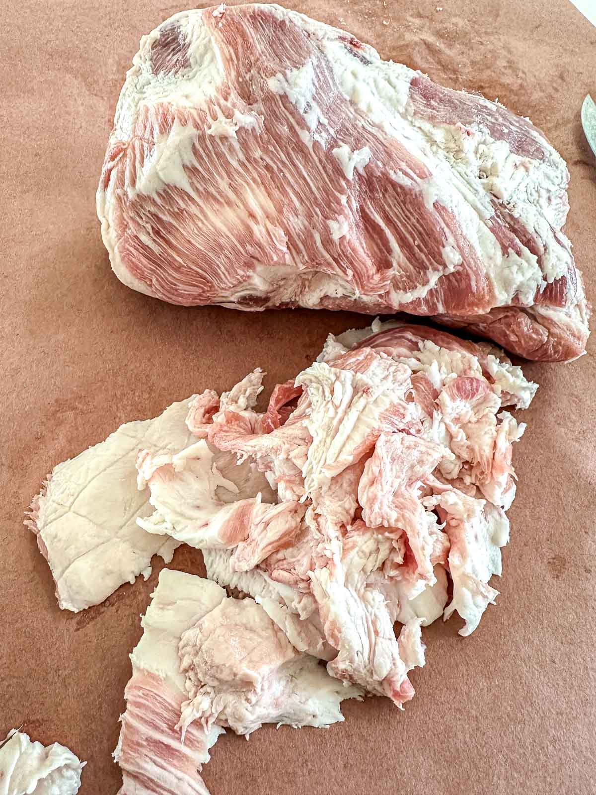 A large piece of raw pork sits on brown paper, with a pile of trimmed fat next to it. A small part of a knife handle is visible in the upper right corner.
