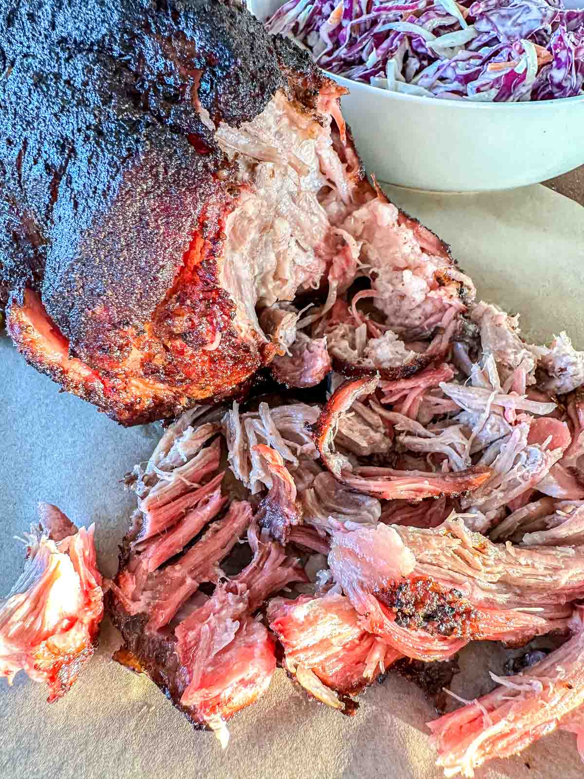 Juicy, shredded smoked pork with a dark, flavorful crust sits on parchment paper. In the background, there is a bowl of creamy coleslaw with purple and white cabbage.