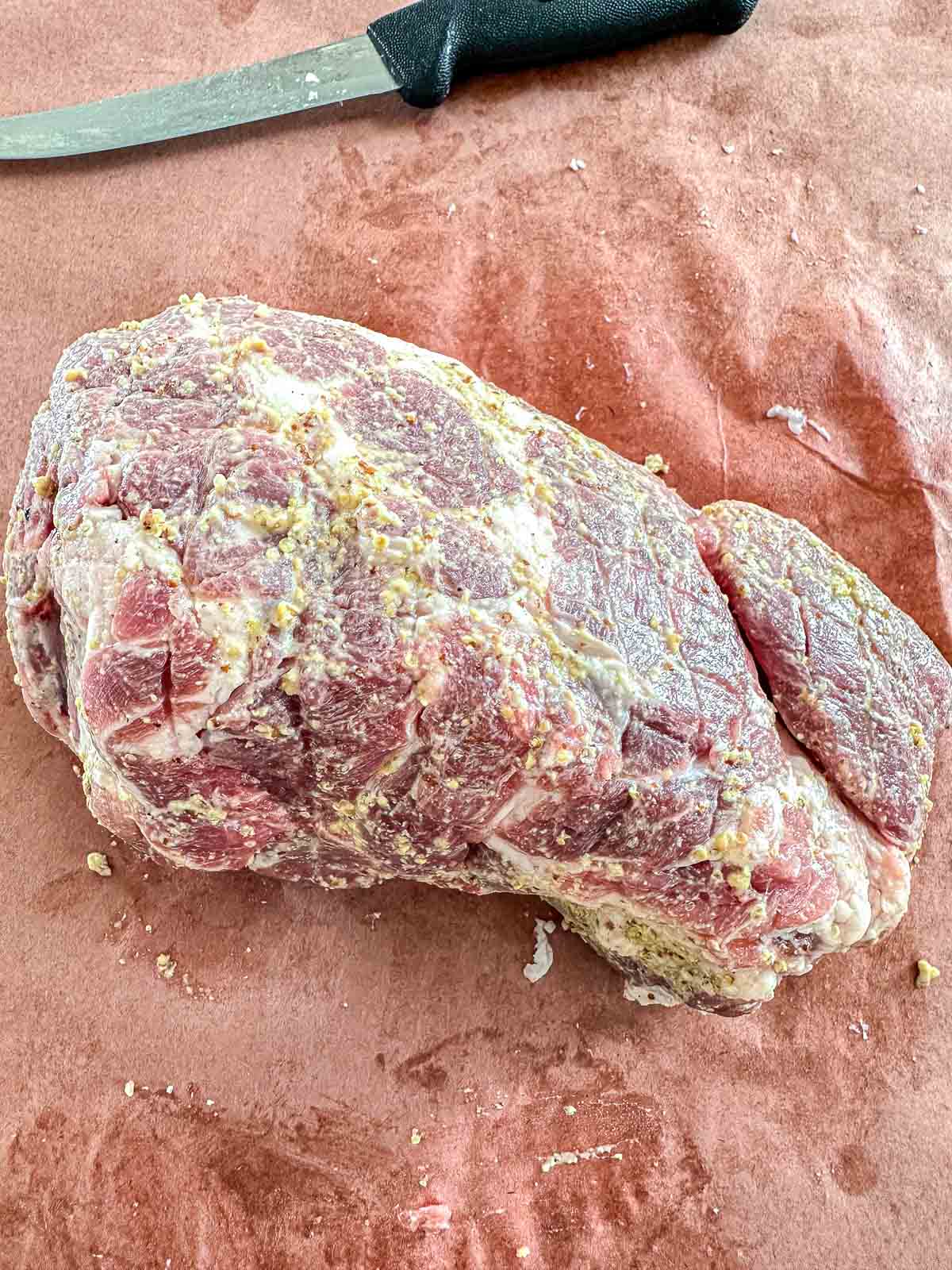 A raw pork roast coated with seasoning sits on brown butcher paper next to a knife with a black handle.