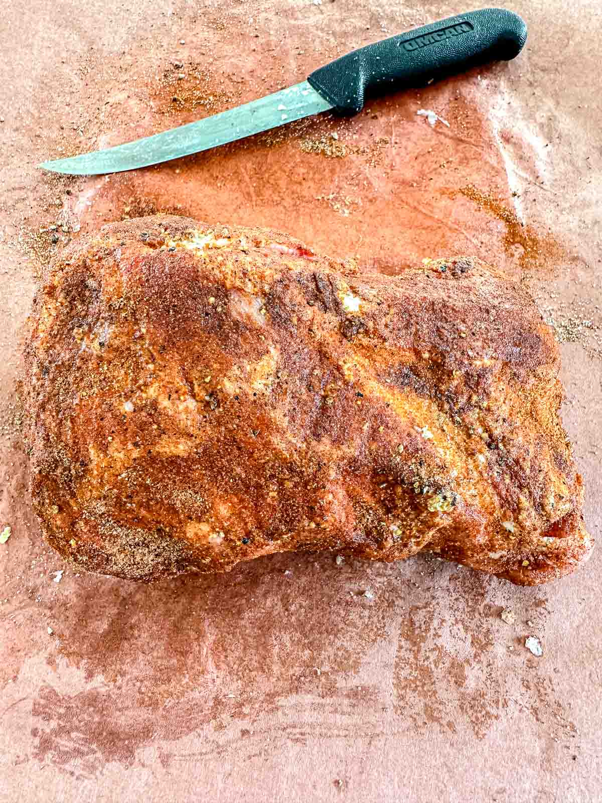 A large, seasoned piece of raw pork butt sits on a brown surface next to a knife with a black handle. The meat is coated with a spice rub.