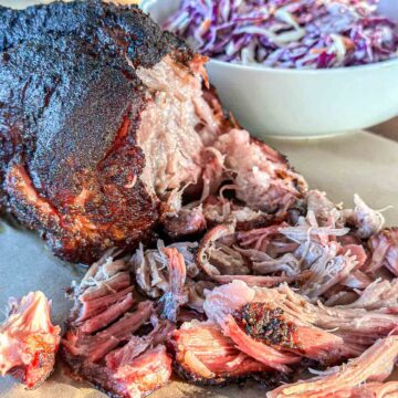 Close-up of smoked, shredded pulled pork with a dark, seasoned crust, served on a cutting board with a bowl of coleslaw in the background.