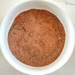 A white bowl filled with a pulled pork rub spice mix sits on a light-colored surface. The seasoning appears finely ground with small, visible specks of different spices.