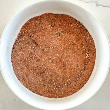 A white bowl filled with a pulled pork rub spice mix sits on a light-colored surface. The seasoning appears finely ground with small, visible specks of different spices.