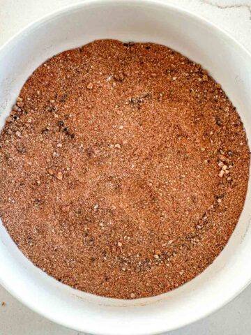 A white bowl filled with a pulled pork rub spice mix sits on a light-colored surface. The seasoning appears finely ground with small, visible specks of different spices.