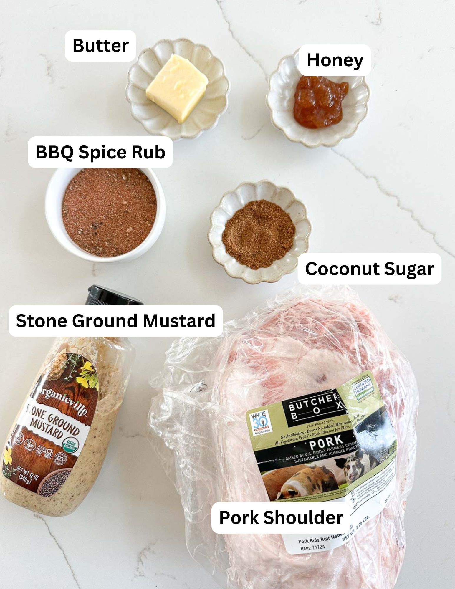 Top-down view of labeled ingredients for a recipe: a wrapped pork shoulder, a jar of stone ground mustard, a small dish of BBQ spice rub, coconut sugar, butter, and honey.