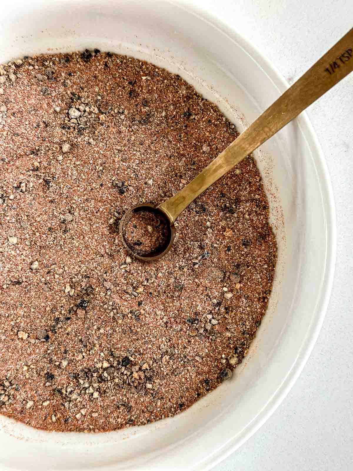 A white bowl filled with a homemade dry rub for smoked chicken, featuring specks of various colors, with a brass ¼ teaspoon resting on top of the mixture.