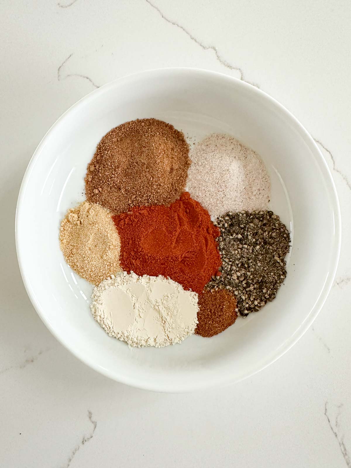 A white bowl containing seven different ground spices arranged in separate piles on a white surface. The spices vary in color, including brown, red, black, beige, and off-white.