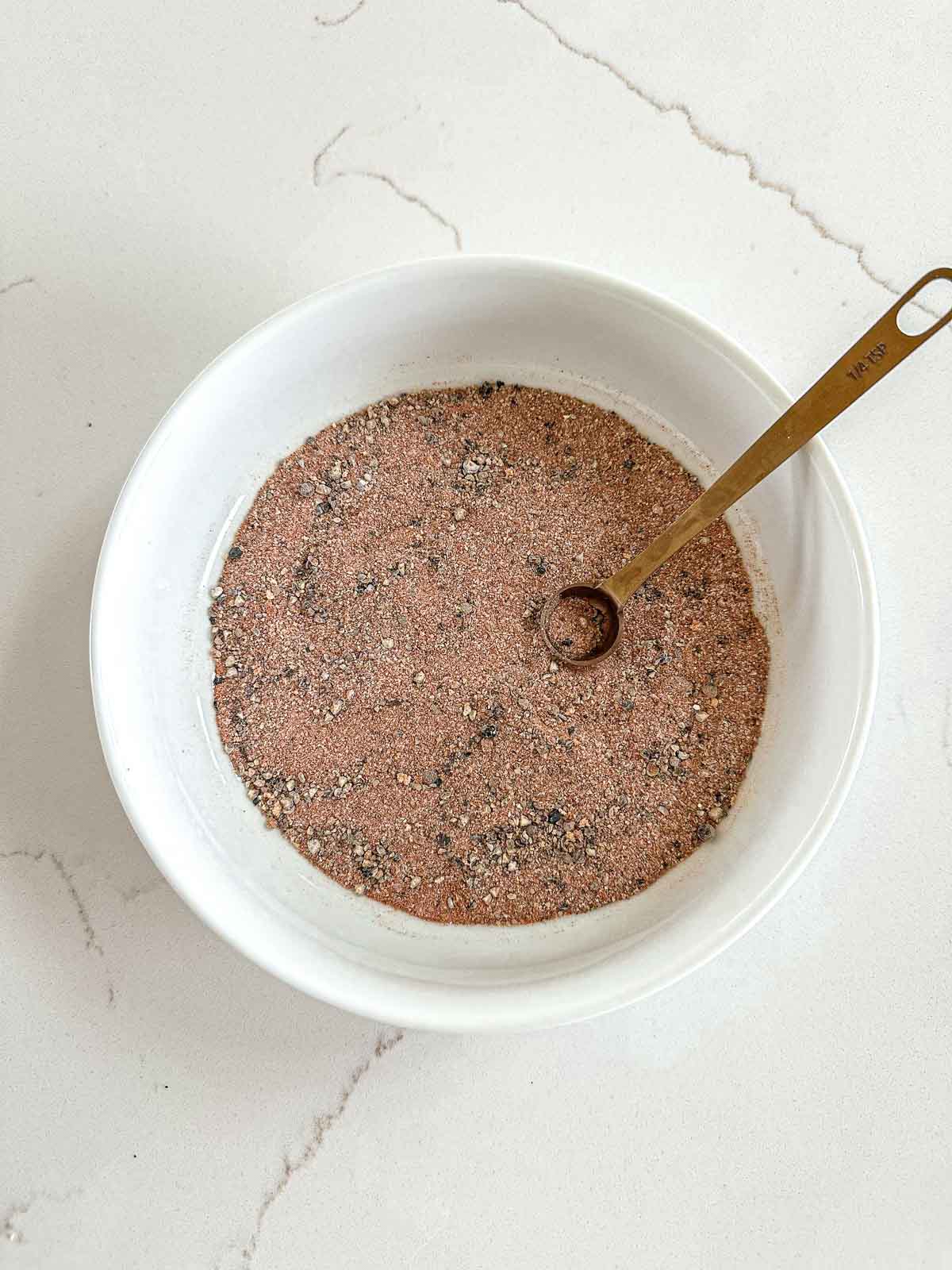 A white bowl filled with a dry rub for smoked chicken sits on a light countertop. A gold measuring spoon rests inside the bowl, partially immersed in the seasoning.