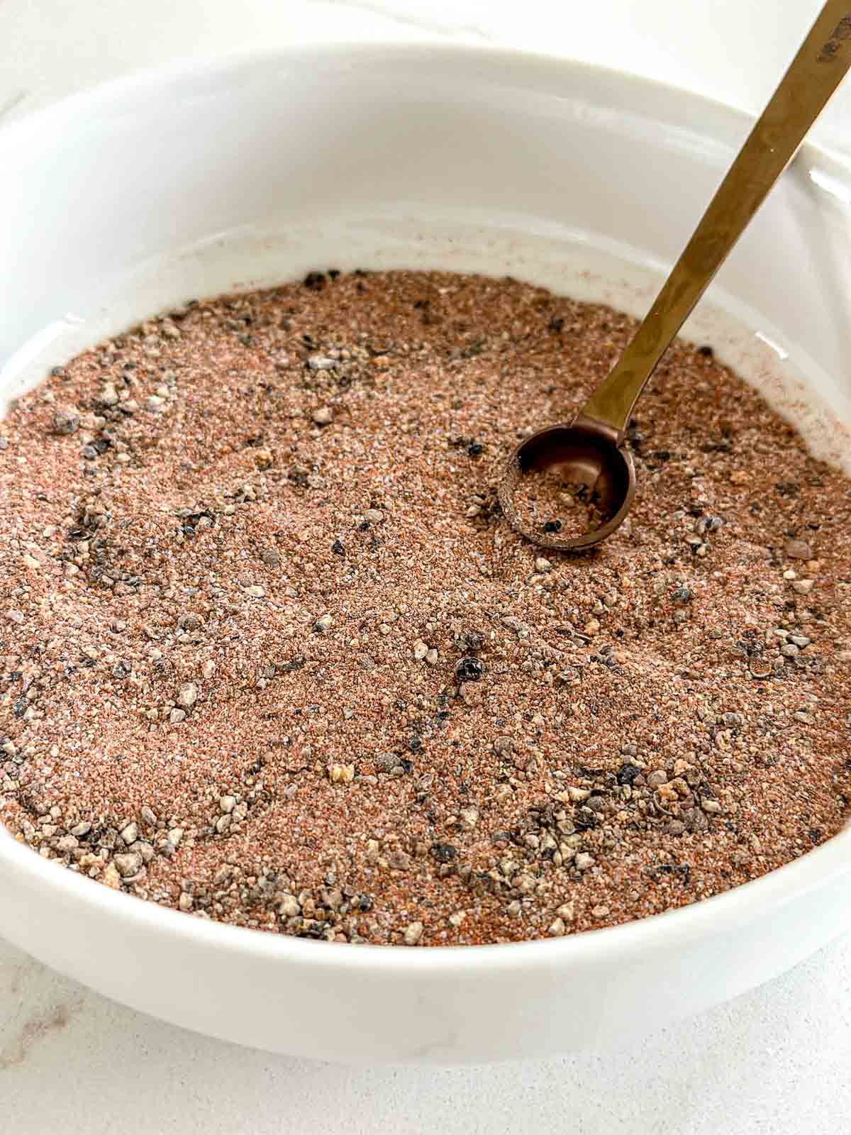 A white bowl filled with a dry rub for smoked chicken and a measuring spoon resting on top.
