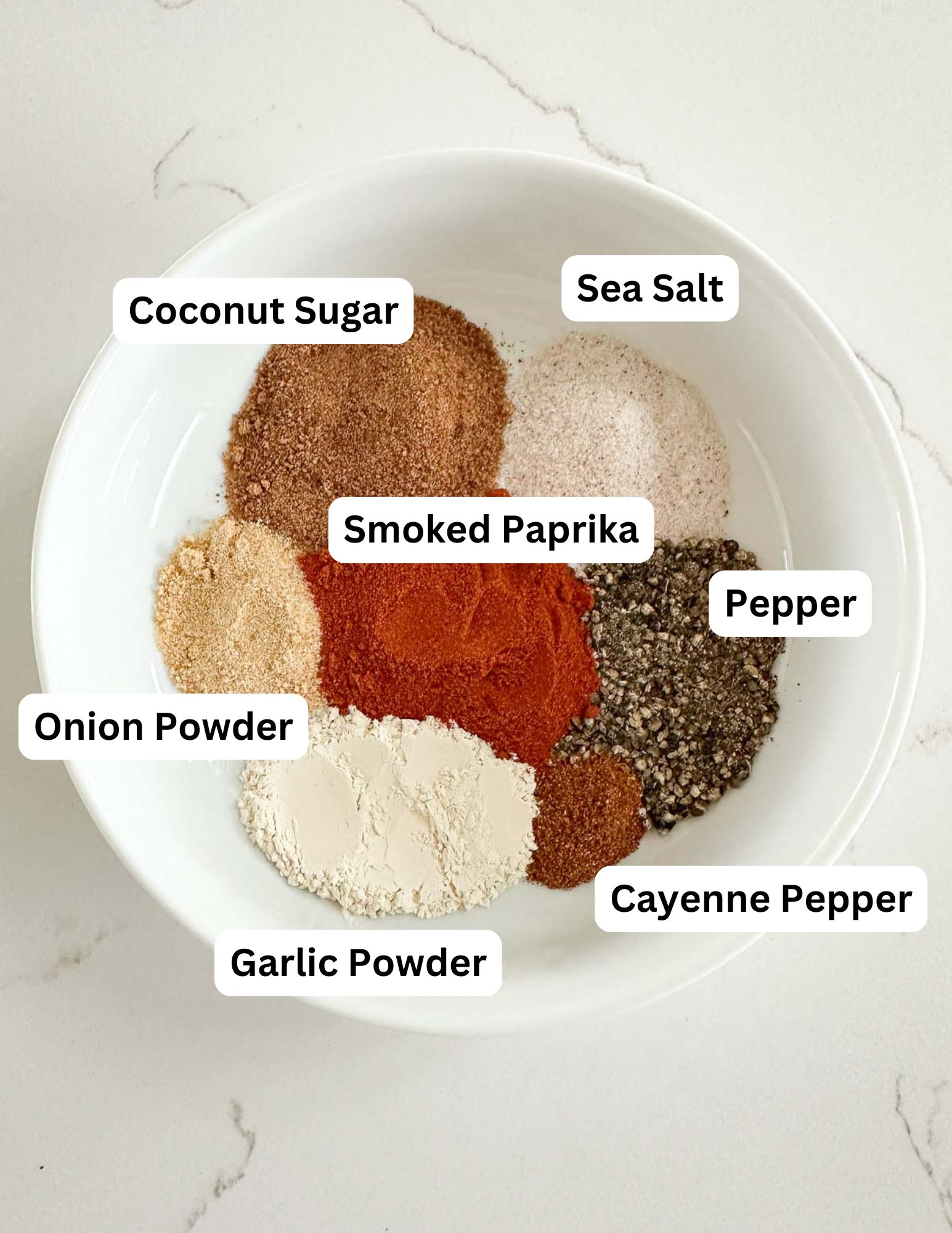 A white bowl contains six labeled spice piles: coconut sugar, sea salt, smoked paprika, pepper, cayenne pepper, garlic powder, and onion powder, set on a light countertop.