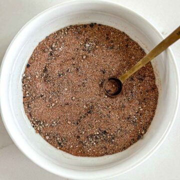 A white bowl filled with a dry rub for smoked chicken with a small metal spoon resting inside. The mixture appears to contain black and brown specks of various textures.
