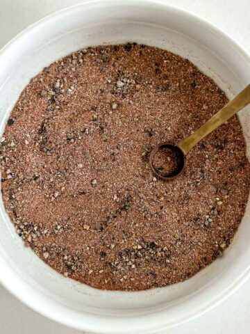 A white bowl filled with a dry rub for smoked chicken with a small metal spoon resting inside. The mixture appears to contain black and brown specks of various textures.