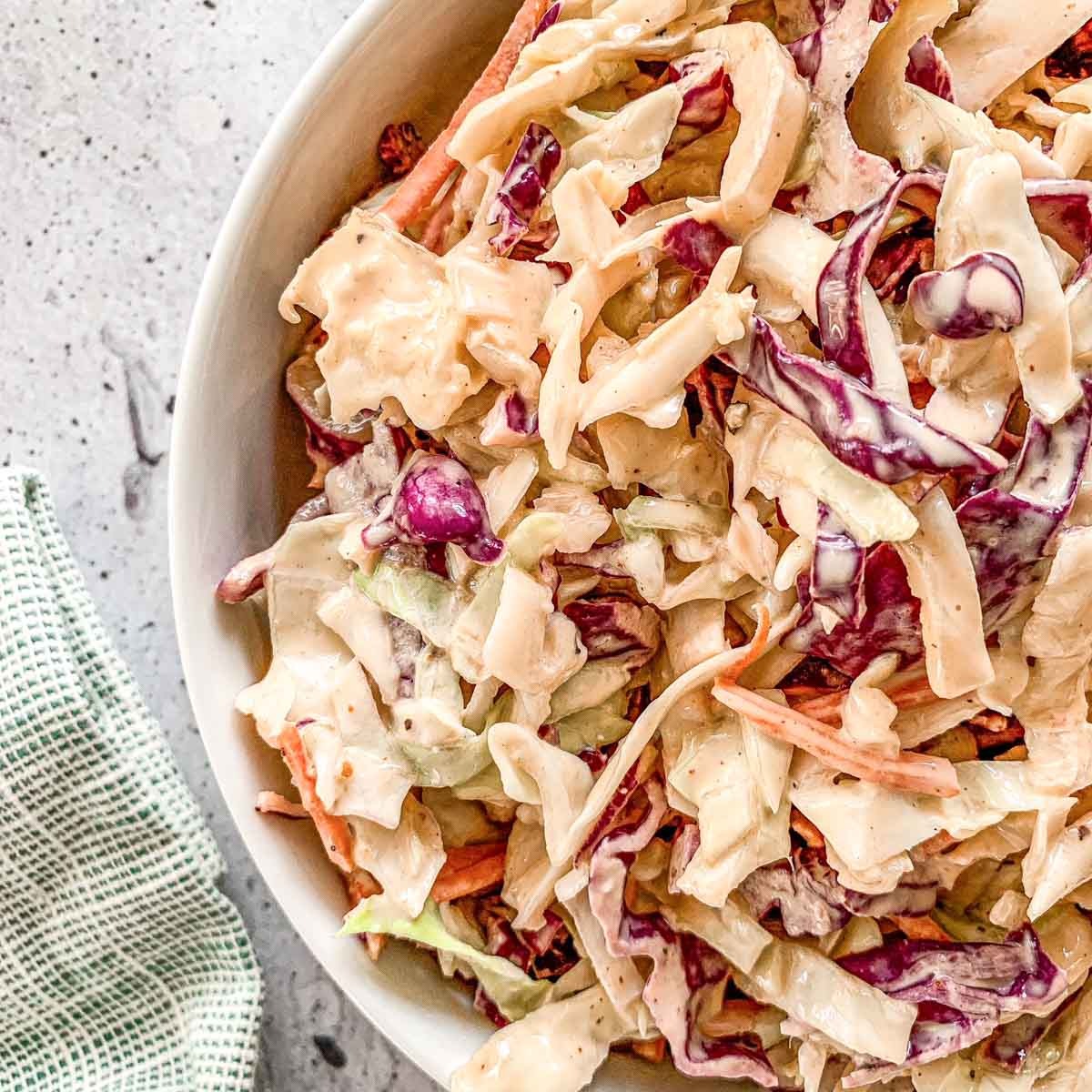 A close-up of a bowl filled with creamy coleslaw made of shredded green and purple cabbage and carrots, set on a light countertop with a green and white checkered cloth nearby.