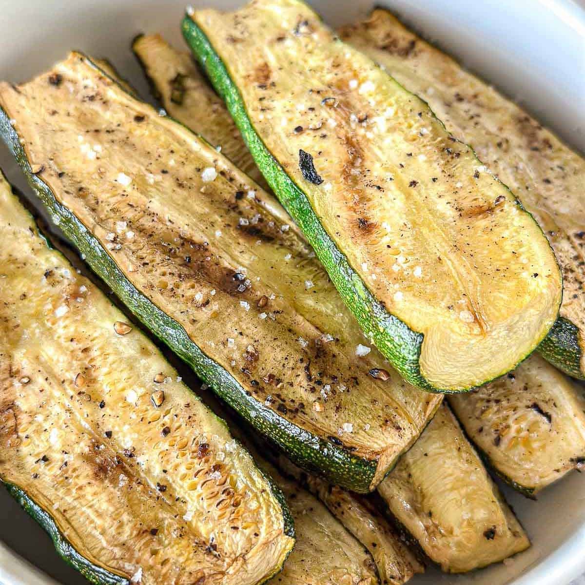 Sliced roasted zucchini seasoned with black pepper and coarse salt, arranged in a white bowl. The zucchini is golden brown with visible grill marks and a slightly crispy texture.
