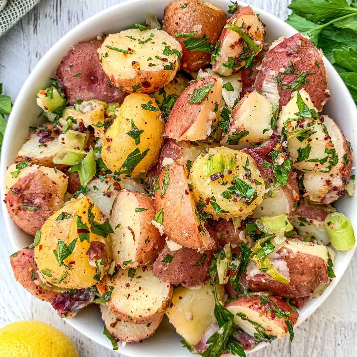 A bowl of potato salad made with red and yellow potatoes, chopped celery, parsley, and seasonings, served on a white surface with lemon and herbs nearby.