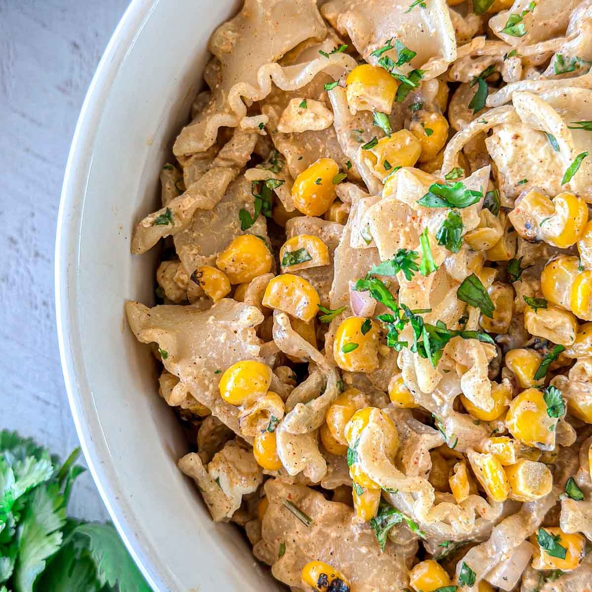 A bowl of pasta salad with roasted corn, chopped herbs, red onion, and a creamy dressing, garnished with fresh cilantro. The dish is served in a white bowl on a light surface.