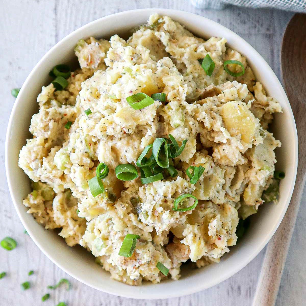 A bowl of creamy potato salad topped with chopped green onions sits on a light wooden surface, with a wooden spoon and scattered green onion slices nearby.
