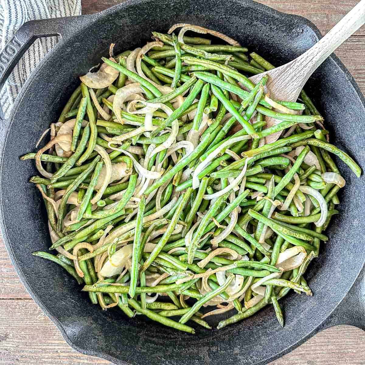 A cast iron skillet filled with sautéed green beans and sliced onions, with a wooden spoon stirring the vegetables. The skillet rests on a wooden surface.