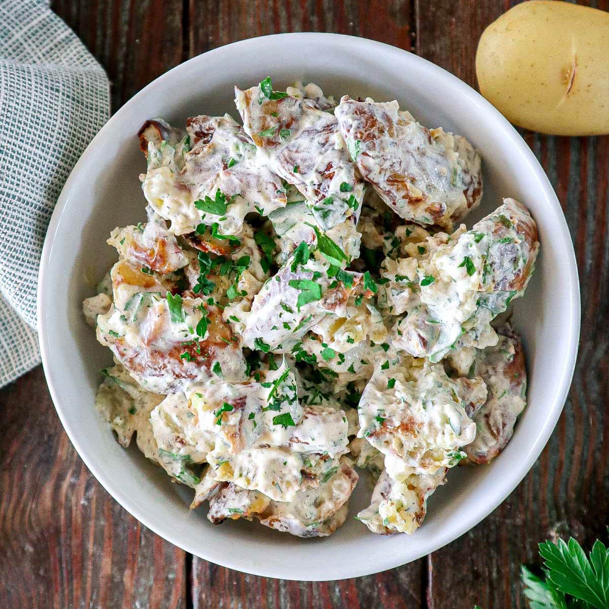 A bowl of creamy potato salad garnished with chopped parsley sits on a rustic wooden table, with a whole potato and part of a cloth napkin nearby.