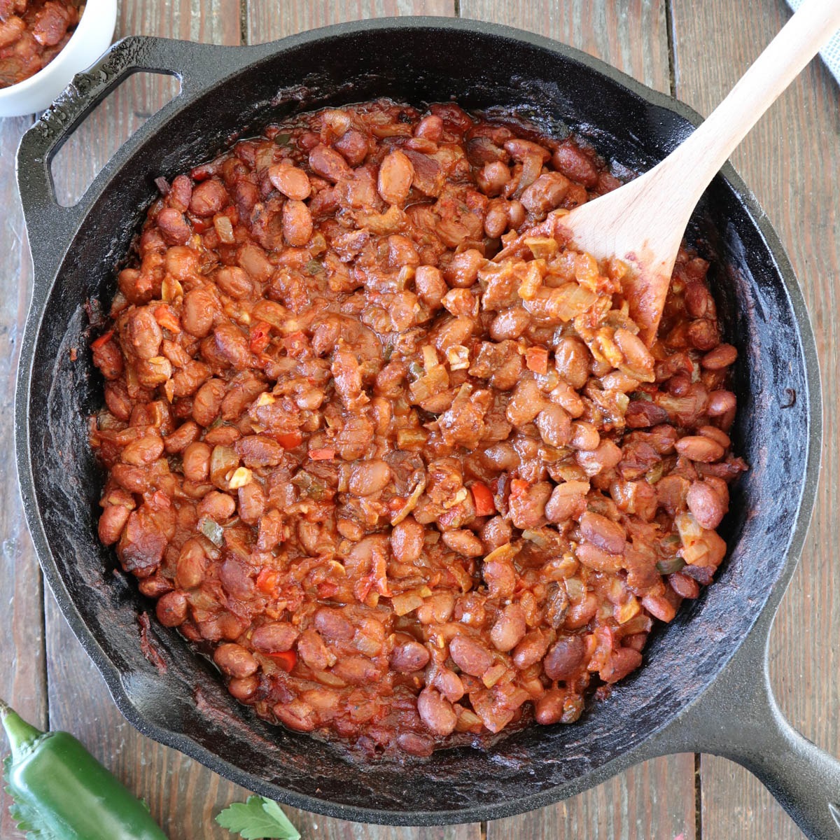 A cast iron skillet filled with baked beans in a thick, reddish sauce, being stirred with a wooden spoon on a rustic wooden table.