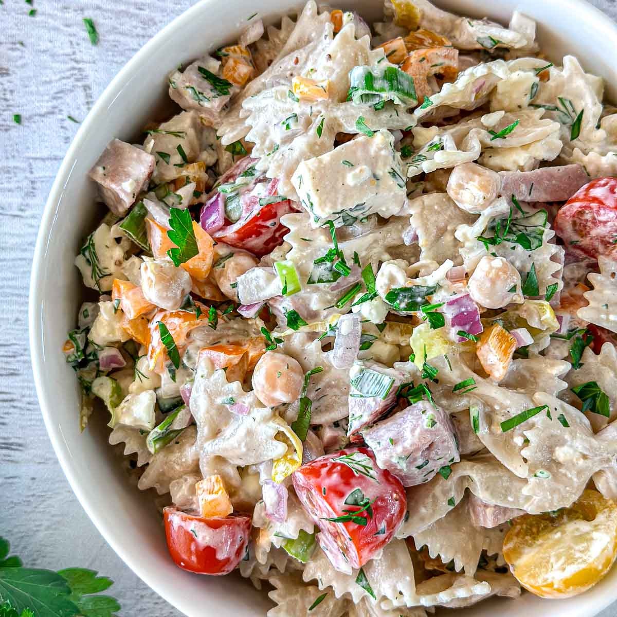 A close-up of a bowl filled with creamy pasta salad, featuring farfalle pasta, cherry tomatoes, chickpeas, diced ham, red onion, bell peppers, and fresh herbs on a light surface.