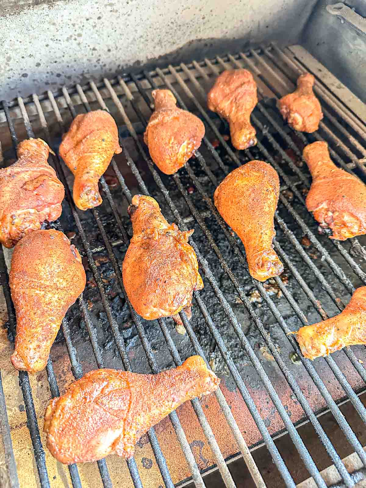 Chicken drumsticks covered in seasoning are cooking on a grill with visible grill marks. The grill is open, showing the metal grates and some char beneath the food.