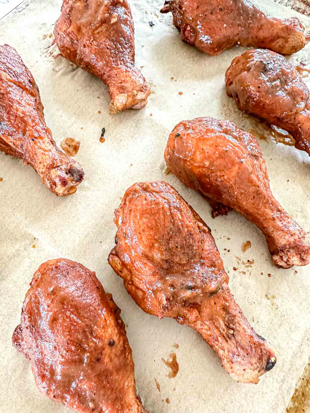 Six cooked chicken drumsticks with a reddish-brown seasoning are arranged on a sheet of parchment paper on a baking tray.