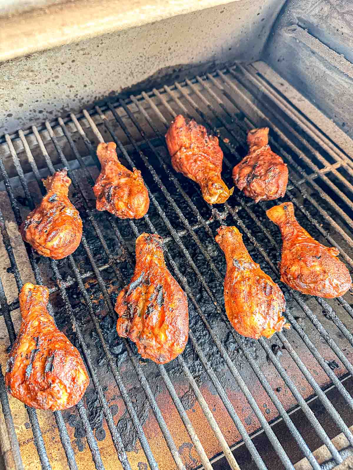 Eight grilled chicken drumsticks with a reddish seasoning are cooking on a barbecue grill, with visible grill marks and some charred spots on the metal grates.