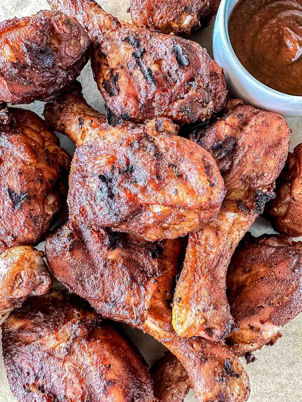 A pile of grilled chicken drumsticks with a smoky, charred appearance sits next to a small bowl of barbecue sauce.
